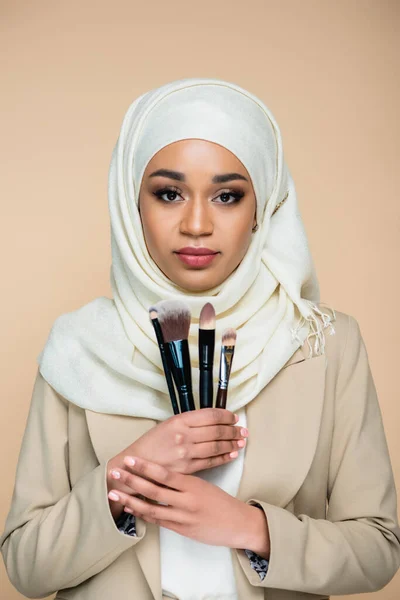 Jovem mulher muçulmana no hijab segurando conjunto de escovas cosméticas isoladas no bege — Fotografia de Stock