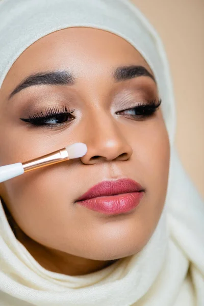Close up of young muslim woman in hijab holding cosmetic brush near face isolated on beige — Stock Photo