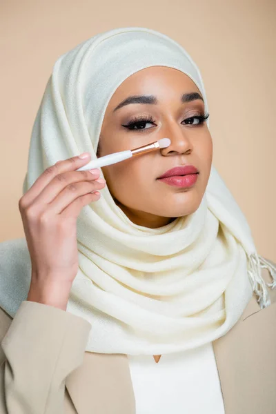 Jovem mulher muçulmana no hijab segurando escova cosmética perto do rosto isolado no bege — Fotografia de Stock