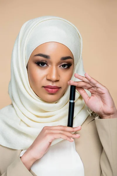 Young muslim woman in hijab holding mascara isolated on beige — Stock Photo