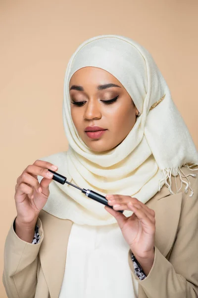 Pretty muslim woman in hijab holding mascara tube and brush isolated on beige — Stock Photo