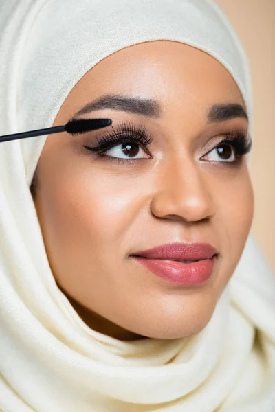 Nahaufnahme einer glücklichen muslimischen Frau im Hijab, die Wimperntusche mit Pinsel isoliert auf beige aufträgt — Stockfoto