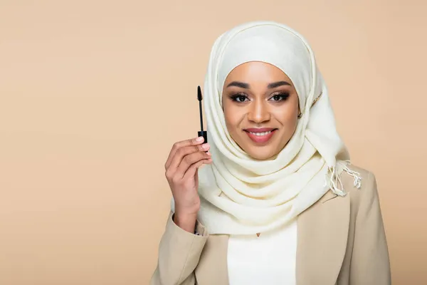 Happy muslim woman in hijab holding mascara brush isolated on beige — Stock Photo