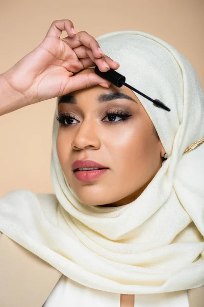 Young muslim woman in hijab holding mascara brush isolated on beige — Stock Photo