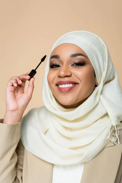 Cheerful muslim woman in hijab holding mascara brush isolated on beige — Stock Photo