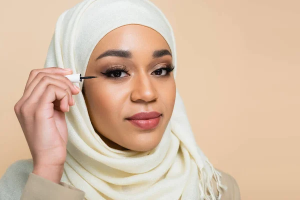 Giovane donna musulmana in hijab applicare fodera occhio nero isolato su beige — Foto stock