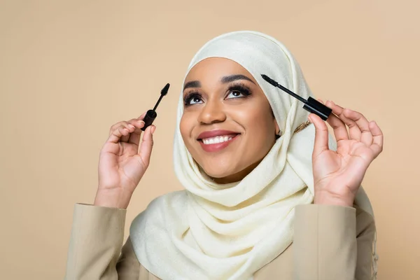 Cheerful muslim woman in hijab holding mascara brushes isolated on beige — Stock Photo
