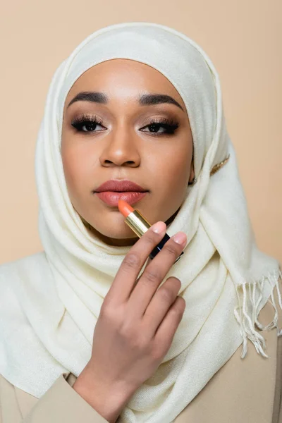 Joven musulmana en hijab aplicando lápiz labial aislado en beige - foto de stock