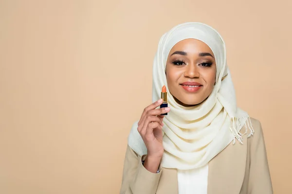 Mujer musulmana feliz en hijab sosteniendo lápiz labial aislado en beige - foto de stock