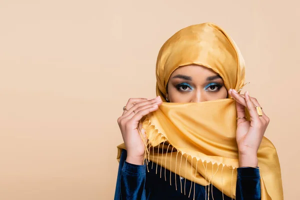 Muslim woman with bright eye makeup covering face with hijab isolated on beige — Stock Photo