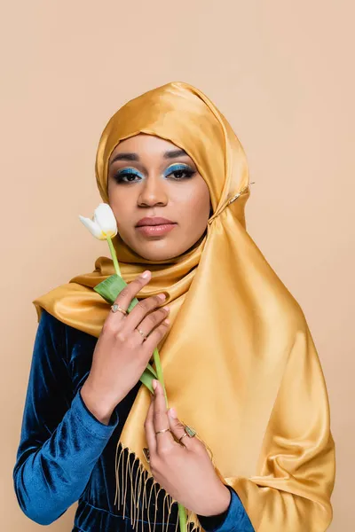 Femme musulmane en hijab avec un maquillage lumineux tenant la tulipe isolée sur beige — Photo de stock