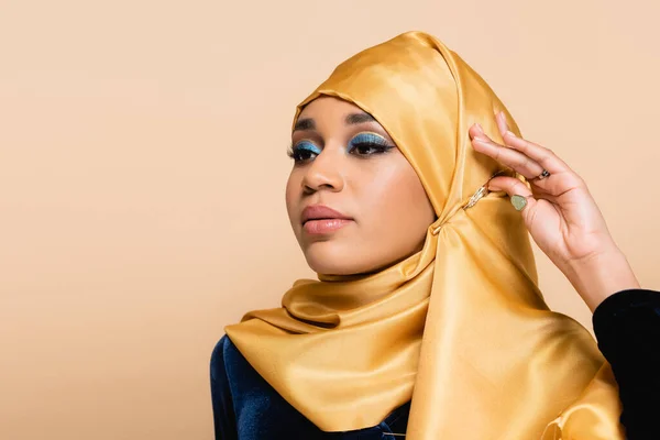 Mujer musulmana joven en hijab amarillo con maquillaje mirando hacia otro lado aislado en beige - foto de stock