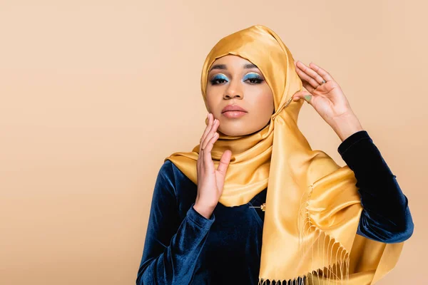 Joven mujer musulmana en hijab amarillo con maquillaje mirando a la cámara mientras posando aislado en beige - foto de stock