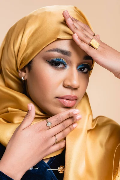 Mujer musulmana en hijab amarillo con maquillaje brillante y anillos de oro mirando a la cámara aislada en beige - foto de stock