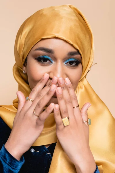 Happy muslim woman in hijab and golden rings covering face while laughing isolated on beige — Stock Photo