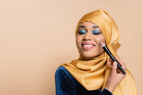 Щаслива мусульманка в жовтому хіджабі наносить макіяж фундаменту з косметичним пензлем ізольовано на бежевому — стокове фото