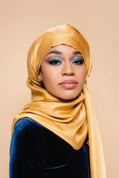 Mujer musulmana en hijab con maquillaje de ojo azul brillante mirando a la cámara aislada en beige - foto de stock