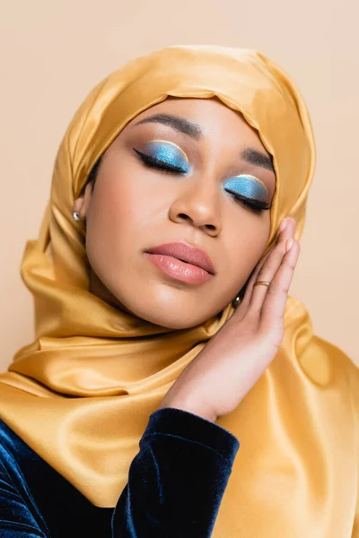 Mujer musulmana en hijab con maquillaje de ojos azul brillante y ojos cerrados aislados en beige - foto de stock