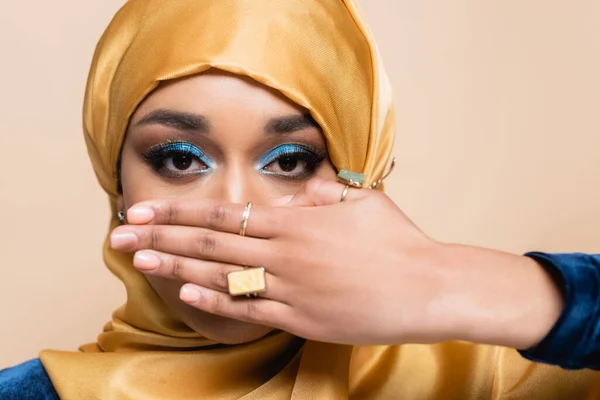 Muslim woman in yellow hijab with golden rings on fingers covering face isolated on beige — Stock Photo