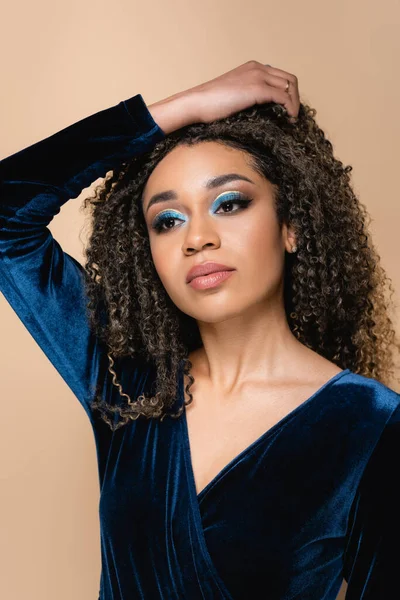 Young african american woman with bright makeup adjusting curly hair isolated on beige — Stock Photo