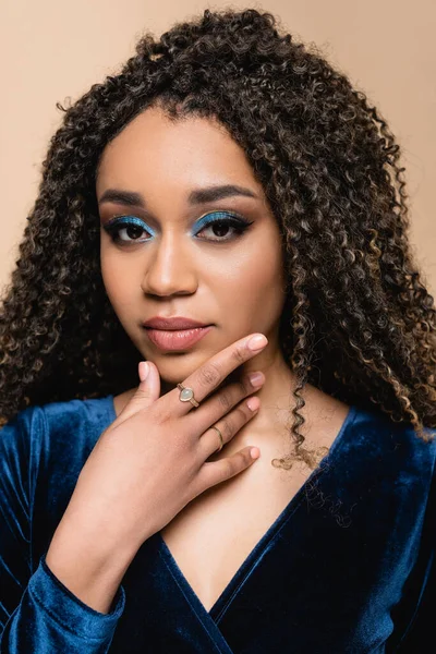 Curly african american woman with makeup touching chin isolated on beige — Stock Photo