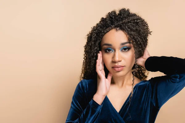 Jolie femme américaine africaine avec un maquillage lumineux ajustant les cheveux bouclés isolés sur beige — Photo de stock