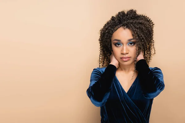 Brunette african american woman with bright blue makeup adjusting curly hair isolated on beige — Stock Photo