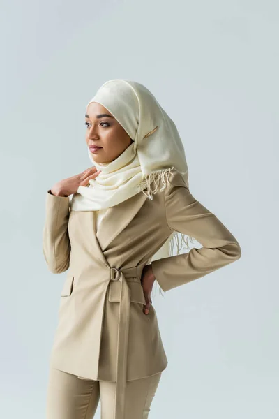 Mujer bastante musulmana en hijab blanco mirando hacia otro lado mientras está de pie con la mano en la cadera aislada en gris - foto de stock