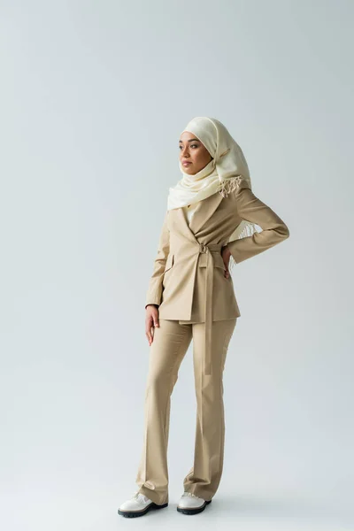 Full length of pretty muslim woman in white hijab and suit standing with hand on hip on grey — Stock Photo