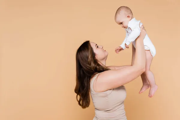 Vista lateral de una mujer sonriente sosteniendo a su hija bebé aislada en beige - foto de stock