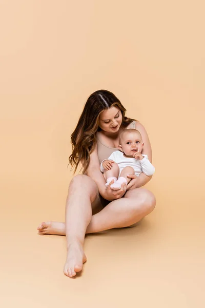 Smiling mother holding baby daughter while sitting on beige background — Stock Photo