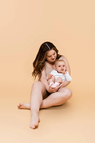 Lächelnde Mutter hält ihre kleine Tochter im Sitzen auf beigem Hintergrund — Stockfoto