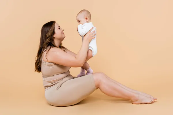 Vue latérale de la mère souriante plus la taille tenant l'enfant tout en étant assis sur fond beige — Photo de stock