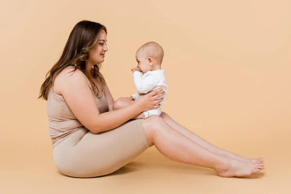 Junge Plus-Size-Frau hält Kind im Sitzen auf beigem Hintergrund — Stockfoto