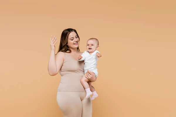 Smiling plus size woman gesturing and holding baby isolated on beige — Stock Photo