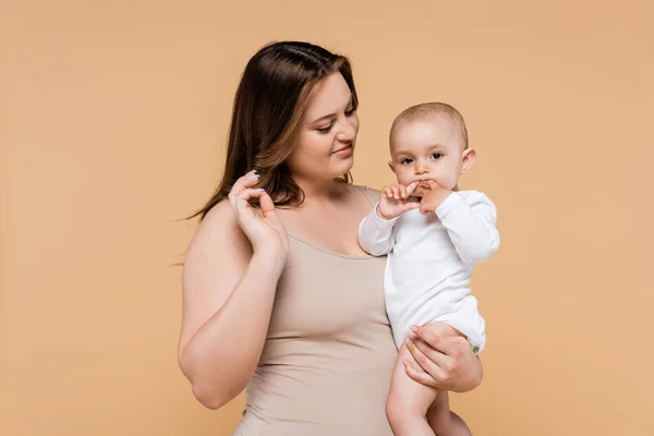 Plus size woman looking at baby daughter holding fingers near mouth isolated on beige — Stock Photo