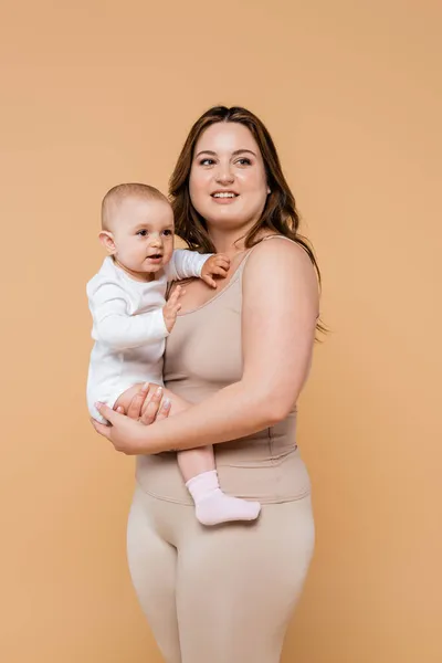 Mujer feliz con sobrepeso sosteniendo al bebé y mirando hacia otro lado aislado en beige - foto de stock