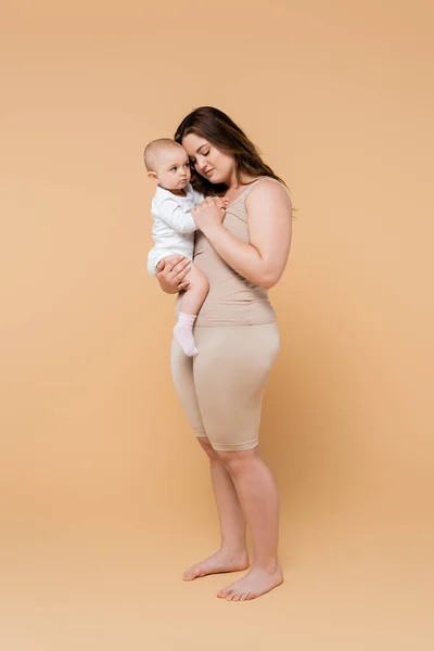 Longitud completa de la madre de talla grande que sostiene a la hija del bebé sobre fondo beige - foto de stock