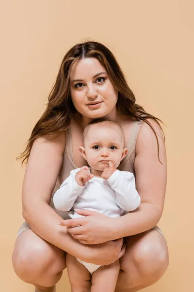Jovem plus size mulher olhando para a câmera enquanto abraça criança isolada no bege — Fotografia de Stock