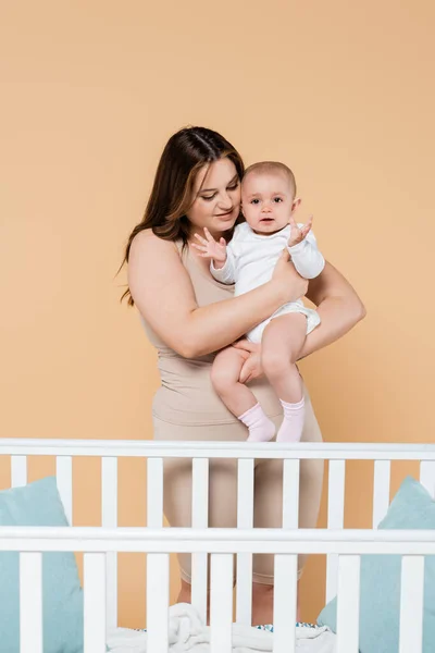 Jeune femme plus la taille tenant bébé fille près du berceau isolé sur beige — Photo de stock