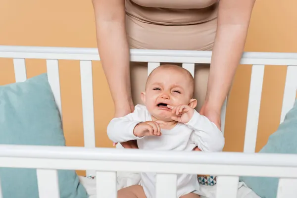 Plus size mother standing near crying baby daughter in crib isolated on beige — Stock Photo