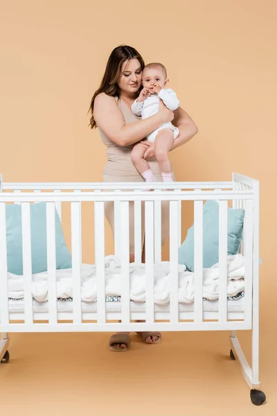 Молодая женщина plus size, держащая ребенка возле кроватки на бежевом фоне — стоковое фото