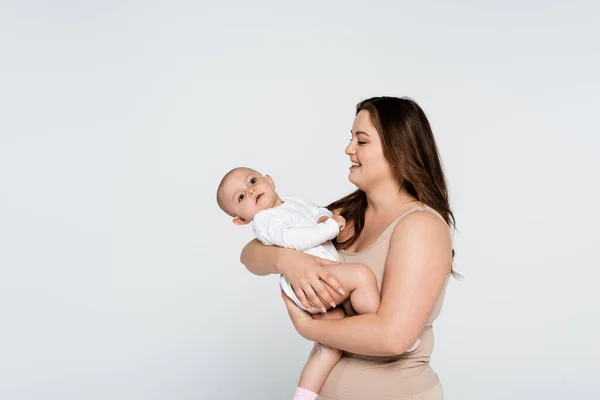 Fröhliche Brünette plus Mutter mit Blick auf Baby-Mädchen isoliert auf grau — Stockfoto
