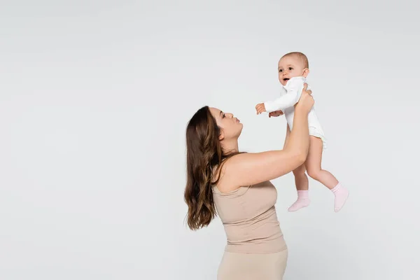 Joven madre con exceso de peso haciendo pucheros mientras levanta a un niño aislado en gris - foto de stock