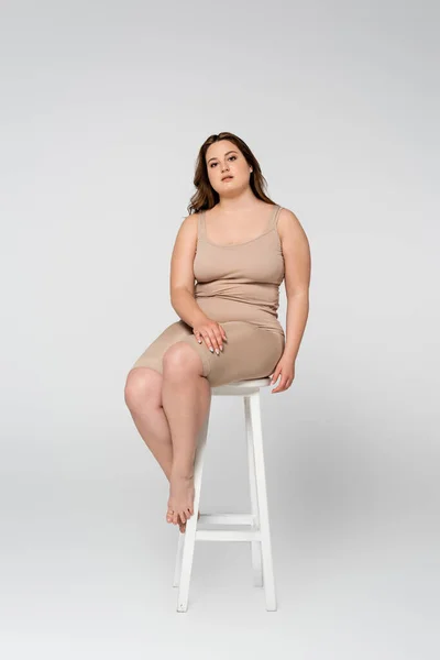 Pretty plus size woman looking at camera while sitting on chair on grey background — Stock Photo