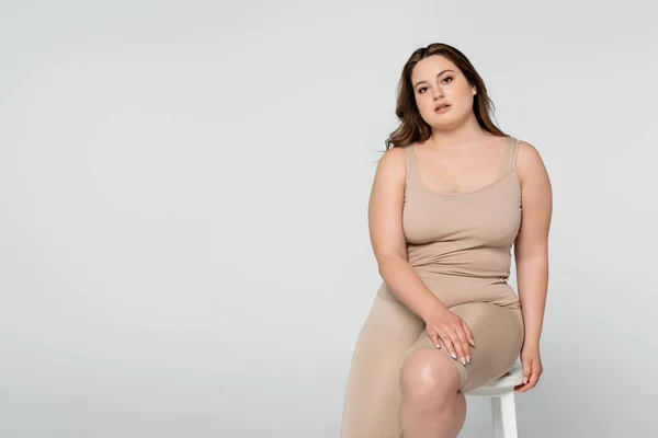 Brunette body positive woman sitting on chair and looking at camera isolated on grey — Stock Photo