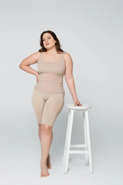 Pleine longueur de jolie femme taille plus tenant la main sur la hanche près de la chaise sur fond gris — Photo de stock