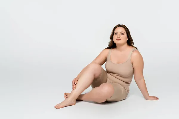 Junge körperpositive Frau blickt in die Kamera, während sie auf grauem Hintergrund sitzt — Stockfoto