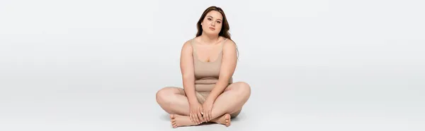 Mujer bonita cuerpo positivo cruzando las piernas mientras está sentado sobre fondo gris, bandera - foto de stock