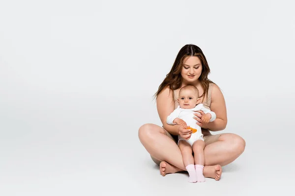 Cheerful body positive woman hugging baby daughter while sitting on grey background — Stock Photo